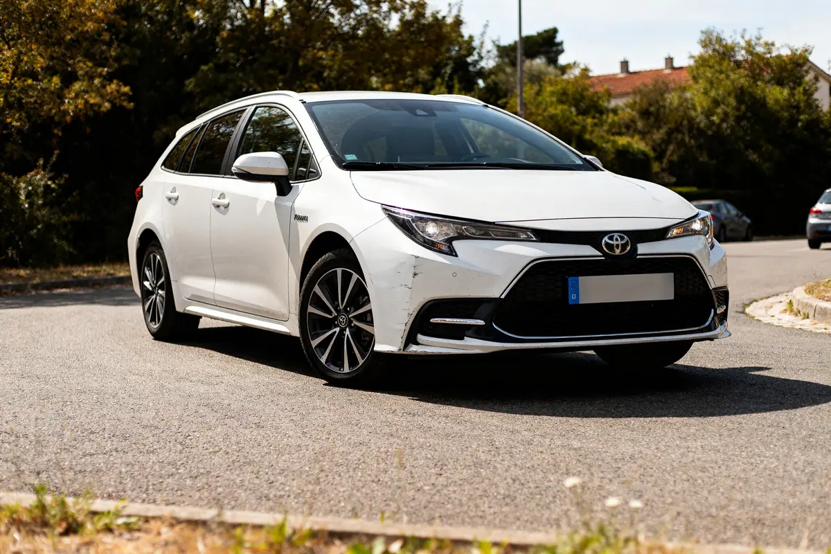 Toyota Corolla Touring blanche stationnée sur route ensoleillée devant arbres et maisons